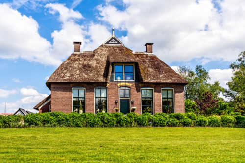 Traditional house with rustic thatched roof in a sunny day. Trees, flowers and bright blue sky with clouds on background. Giethoorn, Netherlands - Starpik