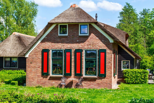 Traditional house with rustic thatched roof in a sunny day. Trees, flowers and bright blue sky with clouds on background. Giethoorn, Netherlands - Starpik