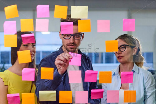 Two caucasian mature women and a man discussing ideas on stickers in the office. View through window - Starpik