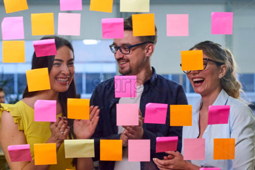 Two caucasian mature women and a man discussing ideas on stickers in the office. View through window - Starpik