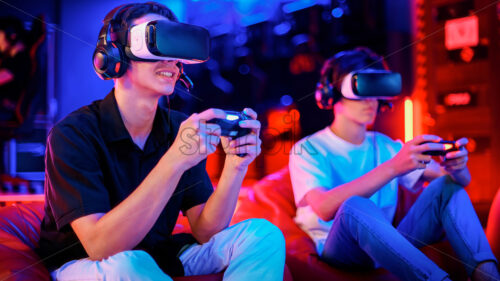 Two teen friends are playing a game console in VR headset and headphones using gamepads and smiling while sitting on bean bags. Blue and red illumination - Starpik