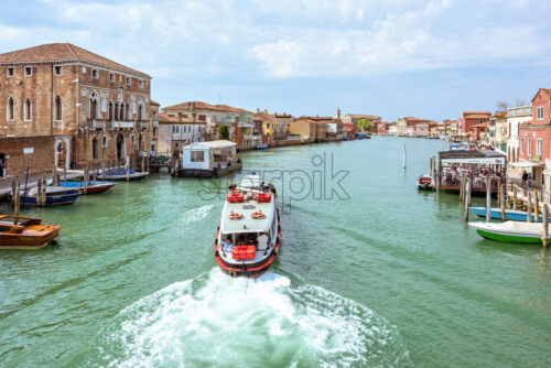 VENICE, ITALY – APRIL 02, 2017: Daylight view to transportation ship cruising to station in Venice Lagoon canal. Parked boats and people walking on sidewalk. Bright blue sky with clouds. Murano Island - Starpik