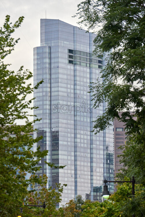 View of the modern skyscraper through the trees with blue cloudy sky behind. The glass facade of this skyscraper reflects the sky - Starpik