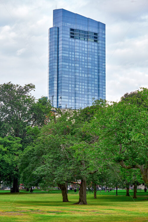 View of the modern skyscraper towering above the park zone with blue cloudy sky behind. The glass facade of the building reflects the sky - Starpik