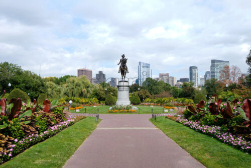 Wide shot of the George Washington Monument located in Boston Public Garden full of greenery with a lot of high modern buildings in the background. The weather is cloudy - Starpik