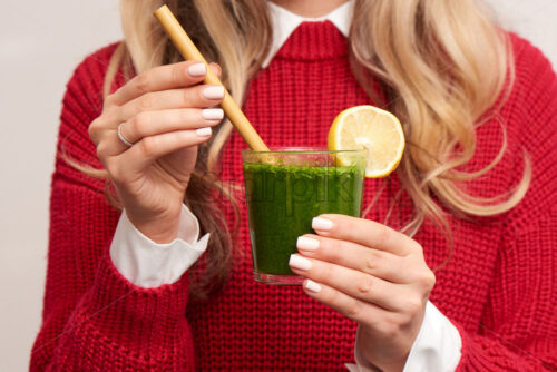 Woman holding fresh spinach green smoothie with lemon decoration and bamboo straw. Red knitted sweater. Healthy products. - Starpik