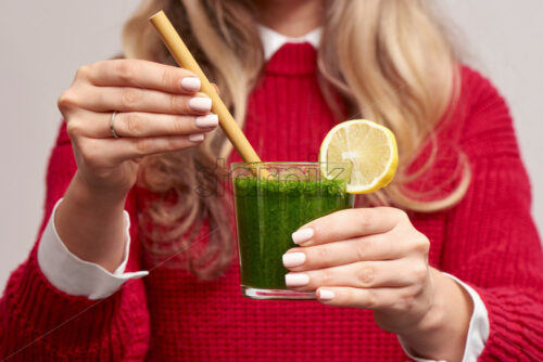 Woman holding fresh spinach green smoothie with lemon decoration and bamboo straw. Red knitted sweater. Healthy products. - Starpik