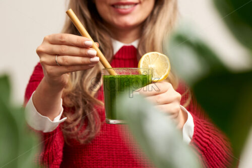 Woman holding fresh spinach green smoothie with lemon decoration and bamboo straw. Red knitted sweater. Healthy products. - Starpik