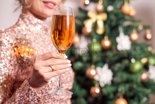 Woman holding glass of champagne with christmas tree on background. Sparkles dress - Starpik