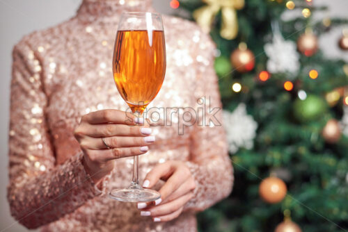 Woman holding glass of champagne with christmas tree on background. Sparkles dress - Starpik