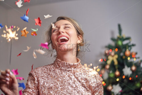 Woman smiling into the camera with christmas tree on background. Holding sparkler. Holiday party. Confetti flying - Starpik