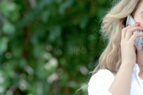 Young blonde woman with pale rose nails talking on smartphone on green trees background. Close-up photo - Starpik