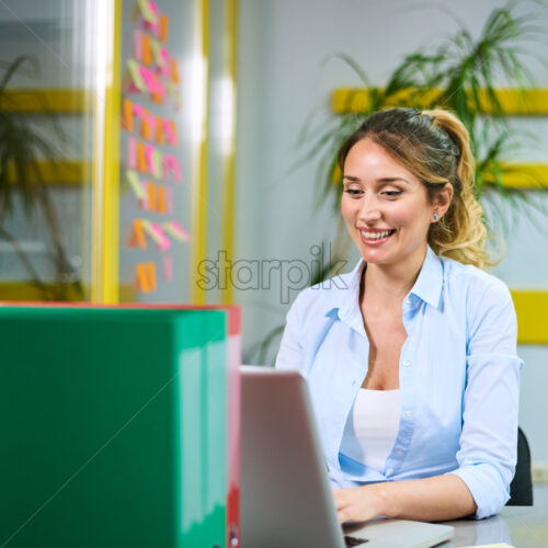 Young business woman working on the laptop computer in the office, smiling and being thoughtful. Workers and sticky notes on background - Starpik