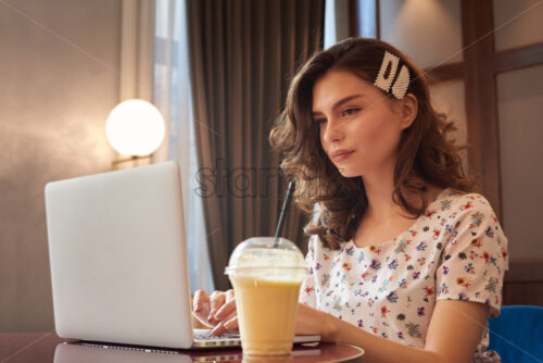 Young cute girl in dress working on a laptop in coffee shop. Milkshake on table. Loft interior - Starpik