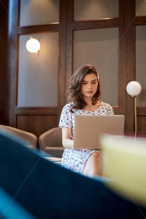 Young cute girl in dress working on a laptop in coffee shop. Sitting on couch. Loft interior - Starpik