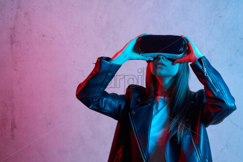 Young girl holding virtual reality helmet while wearing a jacket. Neon red and blue lights on concrete background. Place for text - Starpik