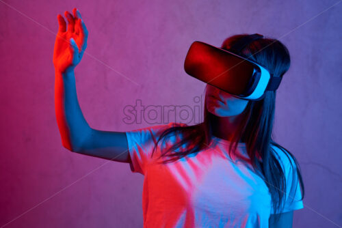 Young girl holding virtual reality helmet while wearing a white t-shirt. Neon red and blue lights on concrete background. Touching with fingers Place for text - Starpik