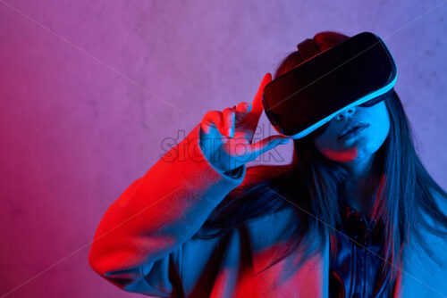 Young girl using virtual reality helmet while wearing a coat. Neon red and blue lights on concrete background. Place for text - Starpik