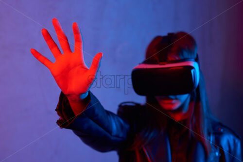 Young girl using virtual reality helmet while wearing a jacket. Neon red and blue lights on concrete background. Holding hand, stop sign. Place for text - Starpik