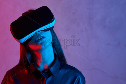 Young girl using virtual reality helmet while wearing a jacket. Neon red and blue lights on concrete background. Place for text - Starpik