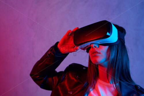 Young girl using virtual reality helmet while wearing a jacket. Neon red and blue lights on concrete background. Place for text - Starpik