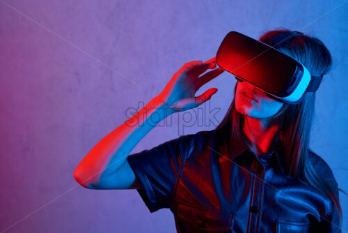 Young girl using virtual reality helmet while wearing a jacket. Neon red and blue lights on concrete background. Touching with fingers. Place for text - Starpik