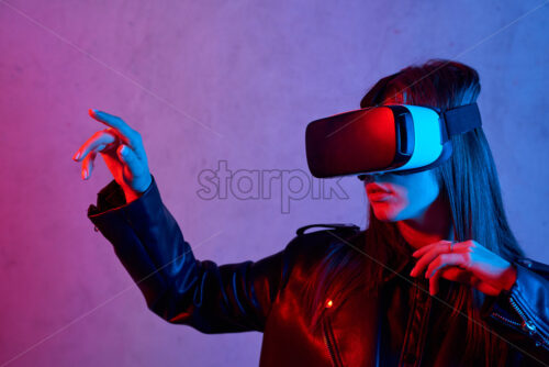 Young girl using virtual reality helmet while wearing a jacket. Neon red and blue lights on concrete background. Touching with fingers. Place for text - Starpik