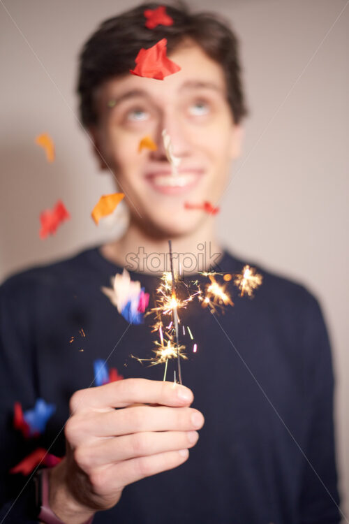 Young man holding sparkler while dreaming about something. Looking up and smiling. Confetti flying. Christmas holiday idea - Starpik