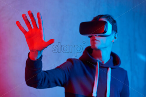 Young man using virtual reality helmet. Neon lights. Wearing sweatshirt. Holding hand in front of red light. View from bottom. Concrete textured wall on background - Starpik