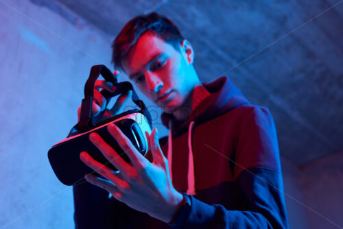 Young man using virtual reality helmet. Neon red and blue lights. Wearing sweatshirt. Preparing to put on head. Concrete textured wall on background - Starpik