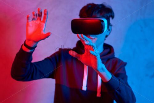 Young man using virtual reality helmet. Neon red and blue lights. Wearing sweatshirt. Scared emotion, holding hands in front of red light Concrete textured wall on background - Starpik