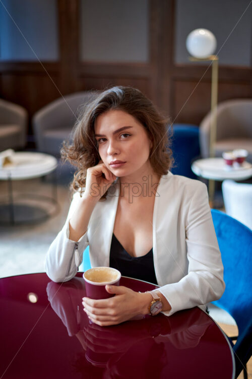 Young sad cute girl drinking coffee in a cafe. Loft interior Looking to the camera - Starpik