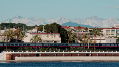 video Train moving in front of buildings and palm trees on the shore with a view of the mountains in the background in Antibes, France Buildings and palm trees on the shore with a view of the mountains in the background in Antibes, France - Starpik Stock